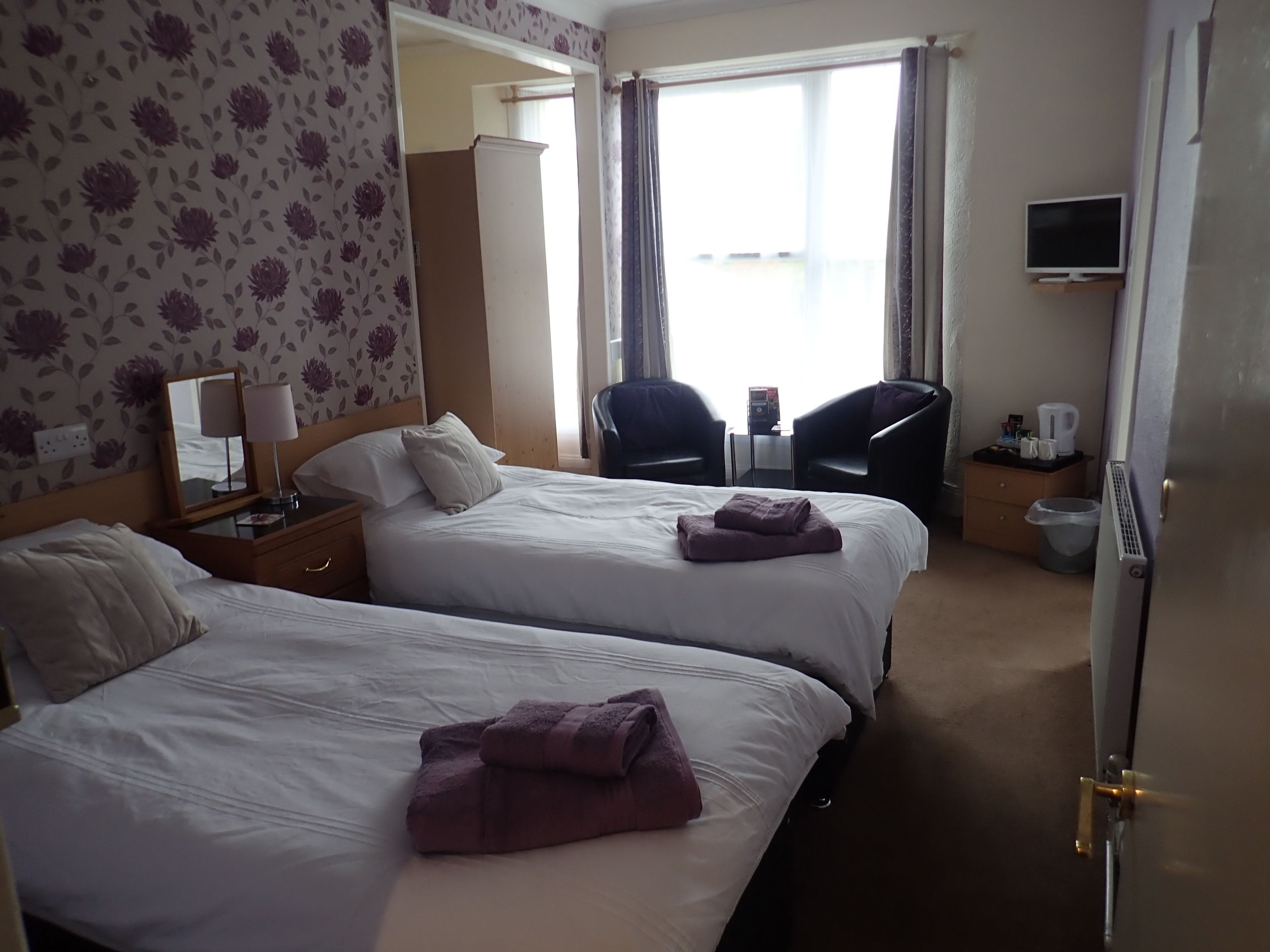 Quarto de hotel com duas camas, uma mesa com abajur, uma cadeira, uma televisão e uma janela com cortinas.