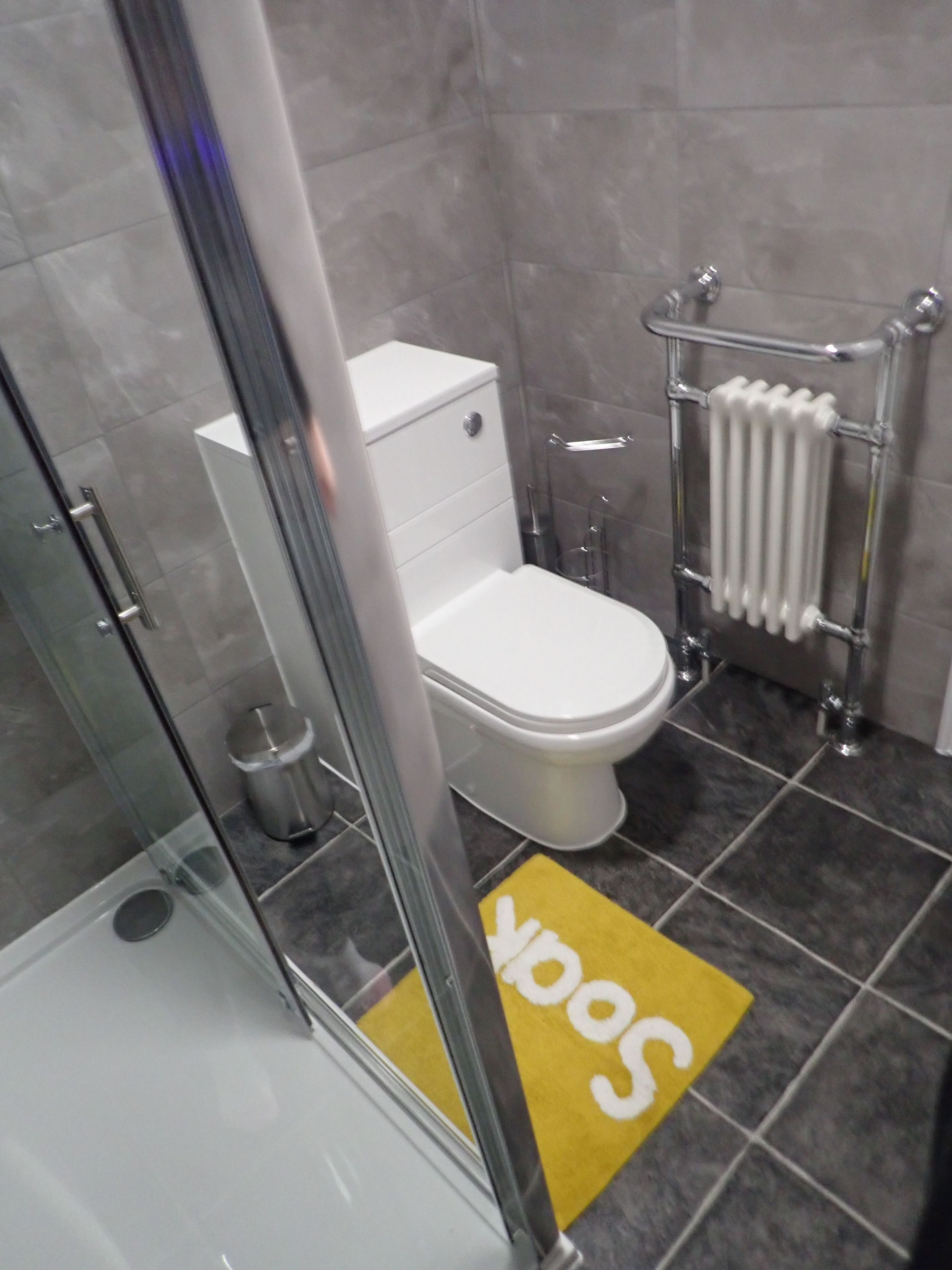 A bathroom with a toilet, a glass shower enclosure, a radiator, and a yellow mat with white text.