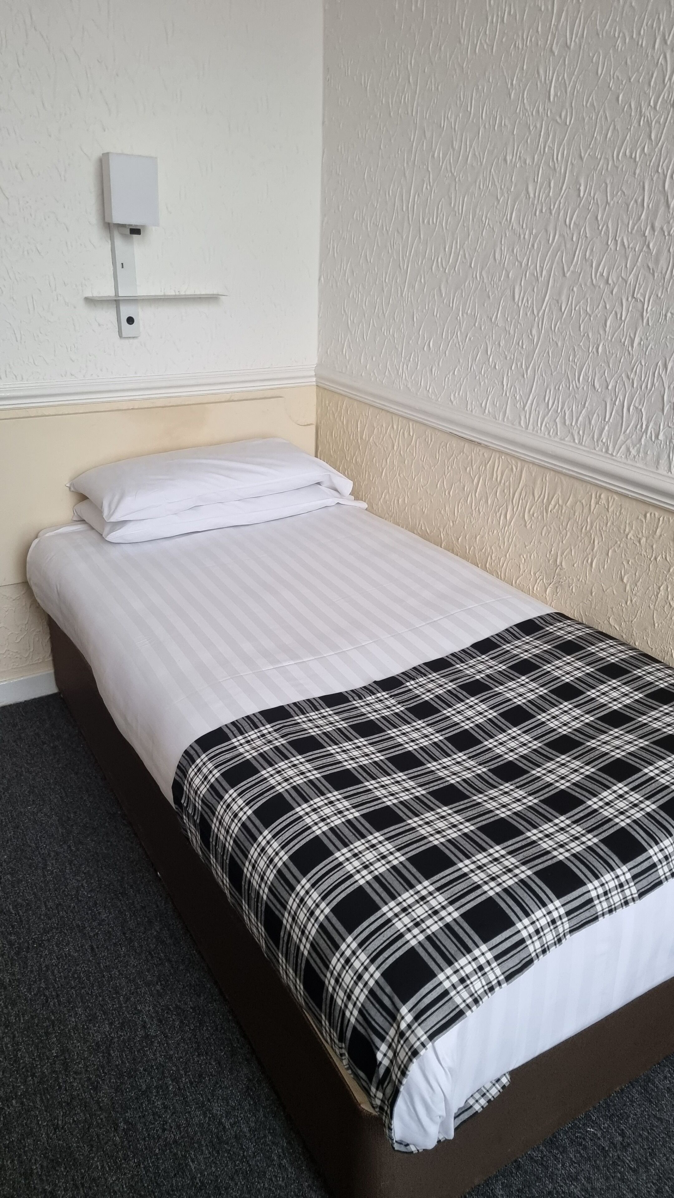 A single bed with a white and black checkered blanket, a white pillow, and a wall-mounted power outlet.