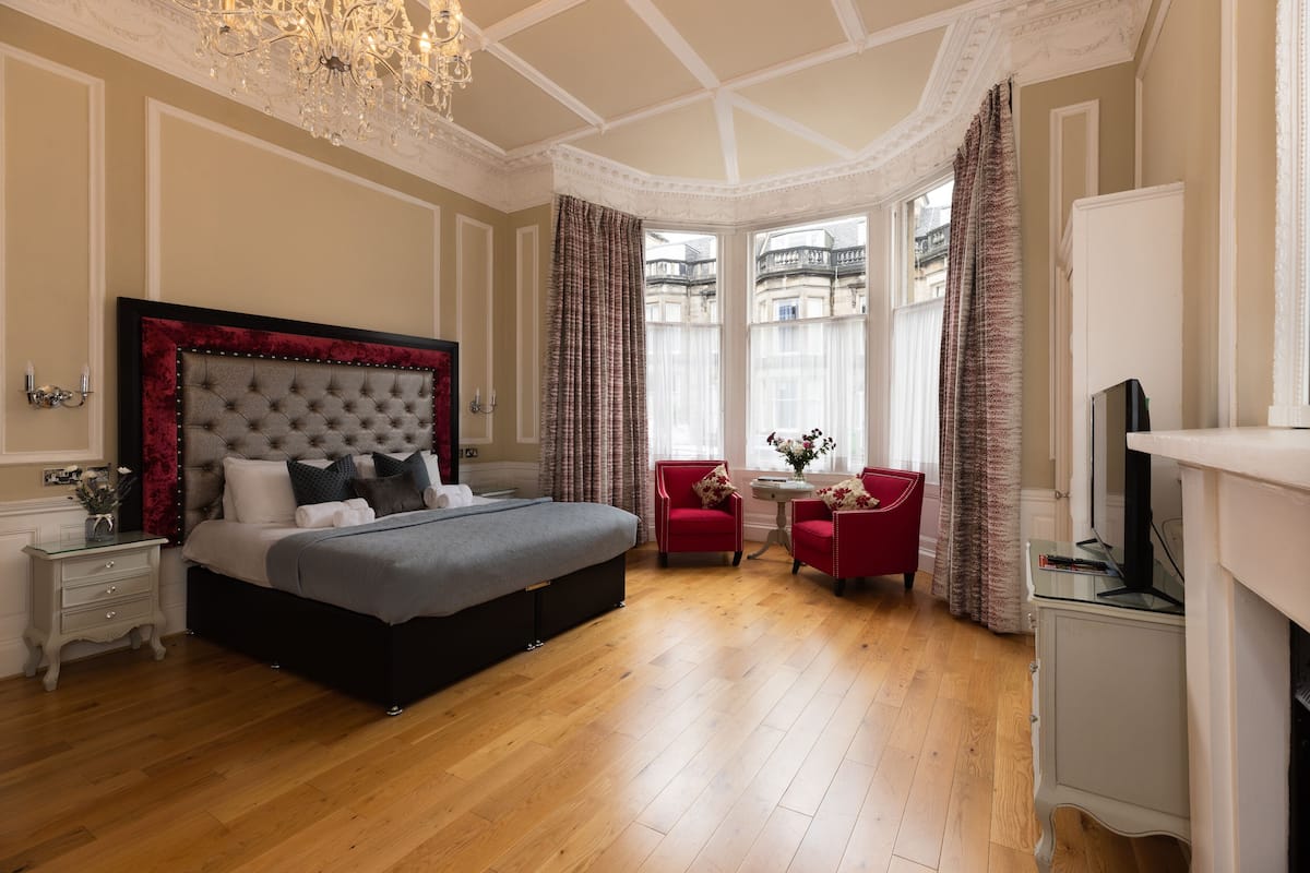 A spacious bedroom with a large bed, a chandelier, a fireplace, and a view of a building through the window.
