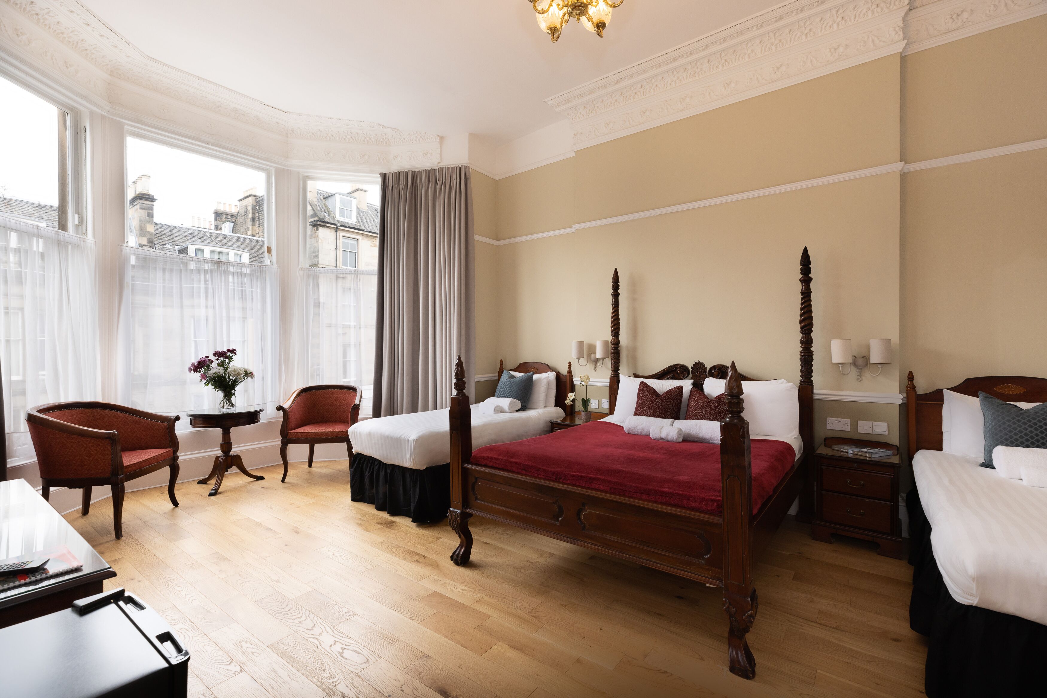A spacious hotel room with a large bed, two chairs, a small table with a vase, and a view of the city through large windows.