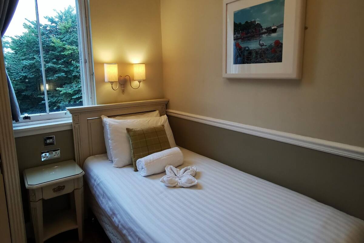 A neatly made bed with a rolled towel and a decorative pillow, a bedside table with a lamp, and a framed picture on the wall.