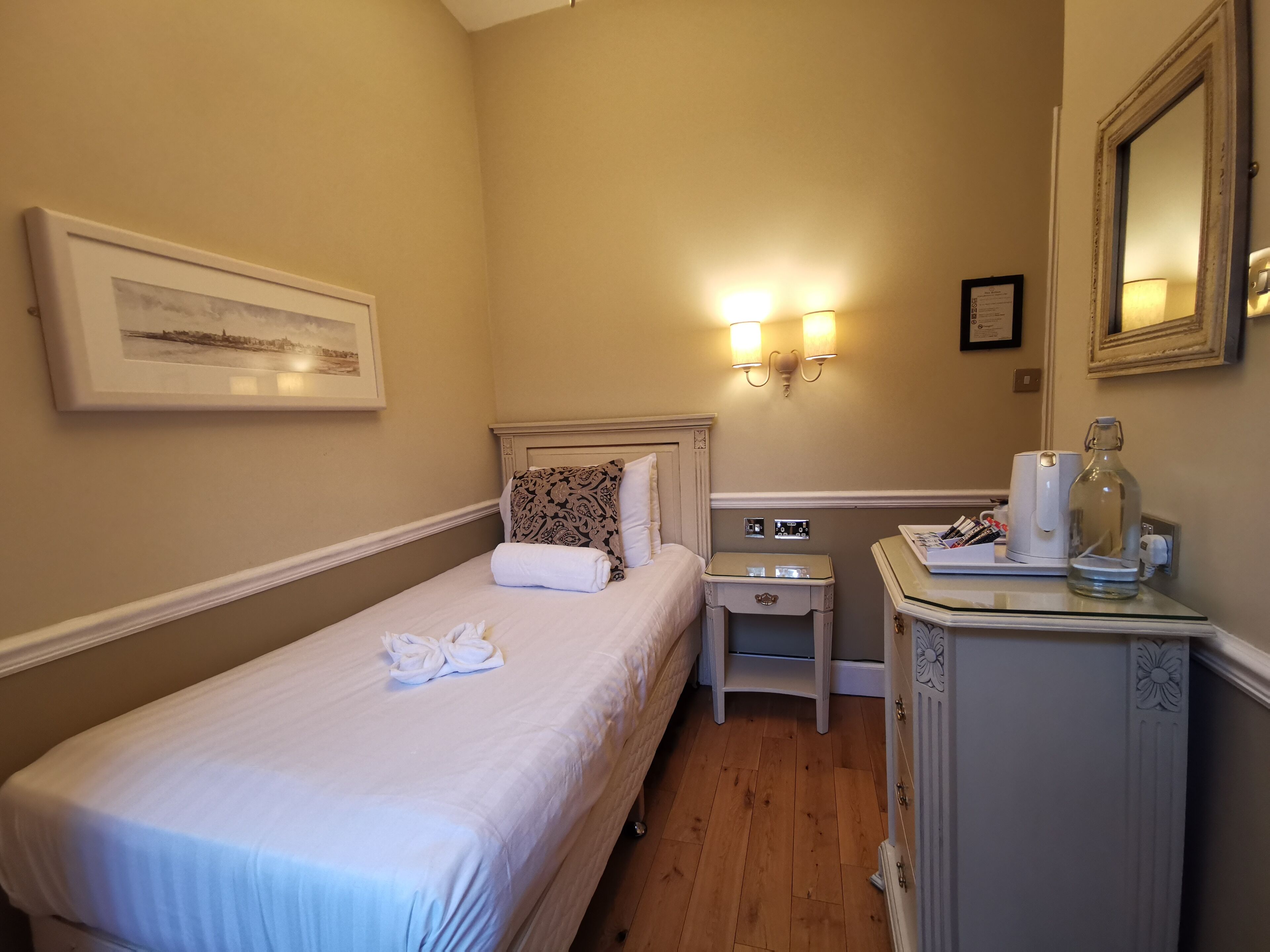 A neatly made bed with a folded towel, a bedside table with a lamp, and a small cabinet with a mirror.