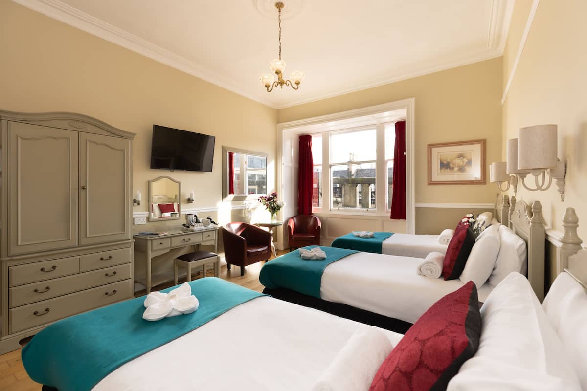 A hotel room with two beds, a desk, a chair, a TV, a window with curtains, and a chandelier.
