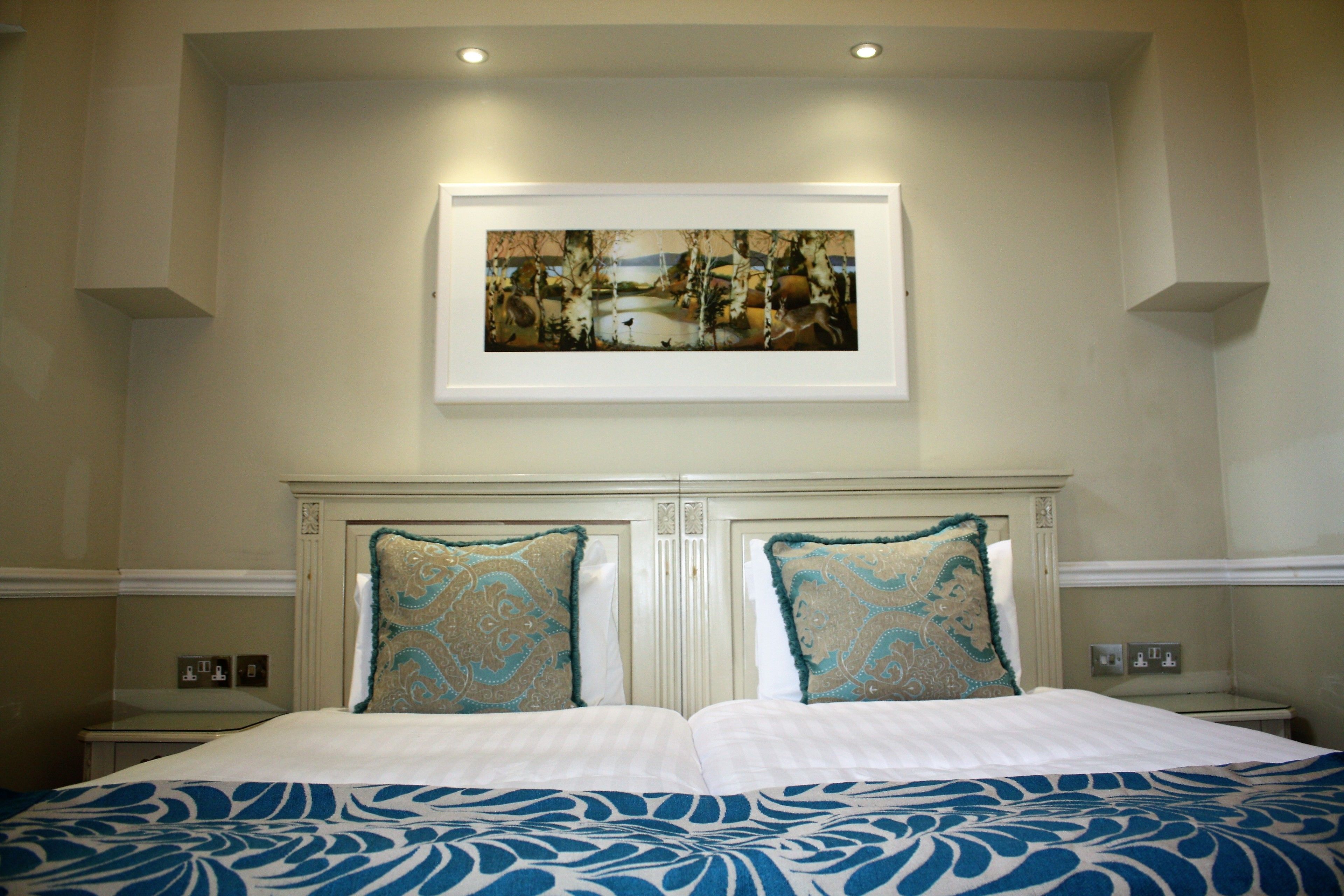 Quarto de hotel com uma cama grande, duas almofadas decorativas, um quadro emoldurado acima da cama e uma mesa de cabeceira com abajur.