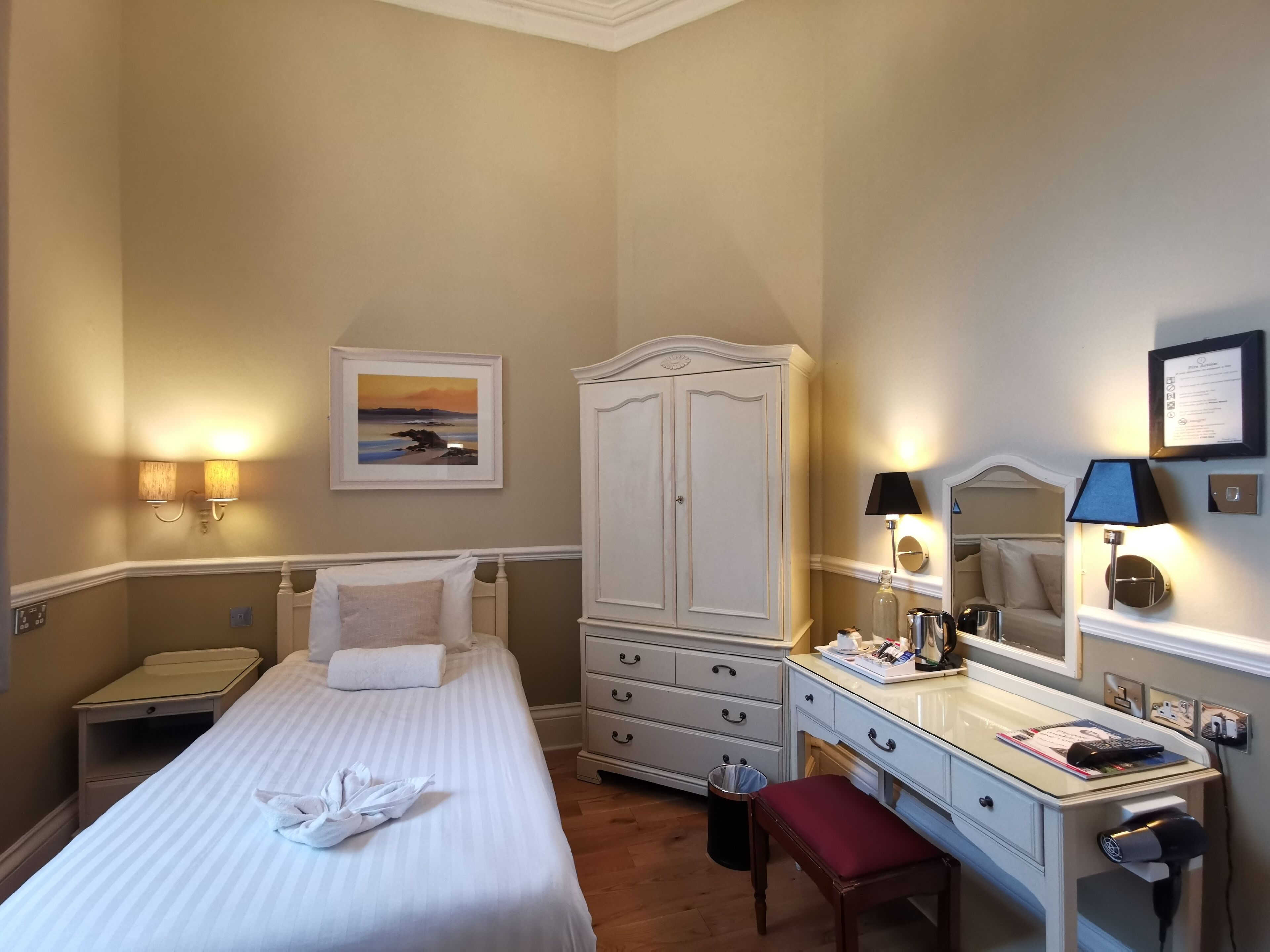 A hotel room with a bed, a desk with a mirror, a wardrobe, and a painting on the wall.