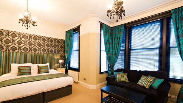 A bedroom with a large bed, a dark sofa, and a coffee table. There are two windows with curtains, a chandelier, and a patterned wallpaper. - The Grafton (Harrogate)