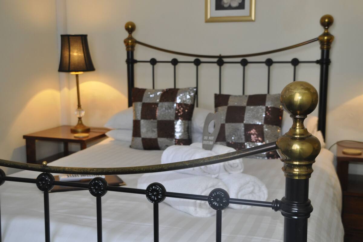 Una cama bien hecha con cabecera metálica, una lámpara de noche y almohadones decorativos.
