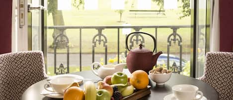 Una mesa de comedor con fruteros, tetera y tazas, ubicada en una habitación con sillas estampadas y vistas a un parque a través de un ventanal.