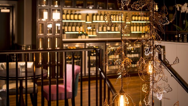 A bar area with a wooden railing, a seating area with a pink chair, and a decorative light fixture with leaf-shaped elements. - West Park Hotel (Harrogate)
