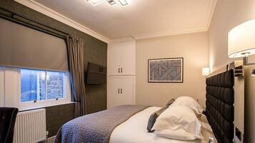 A bedroom with a bed, a bedside table, a lamp, a radiator, a window with curtains, and a wall-mounted artwork.