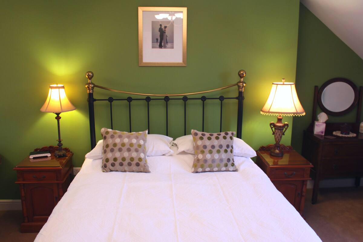 A neatly made bed with two pillows, two bedside tables with lamps, a framed picture on the wall, and a small wooden nightstand with a mirror and a tissue box.