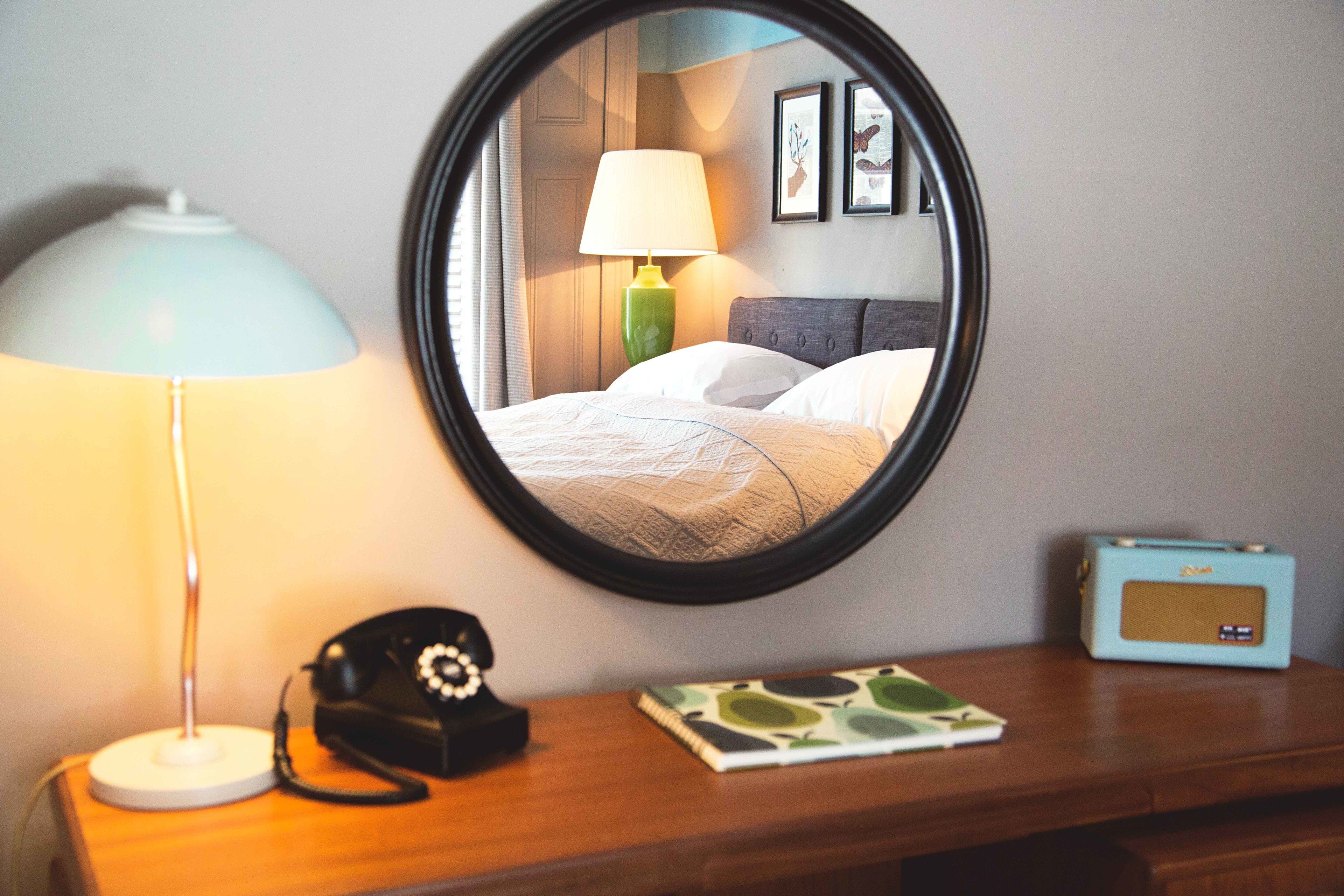 Un espejo redondo refleja una cama, una lámpara, una radio y un teléfono sobre una mesita de noche de madera.