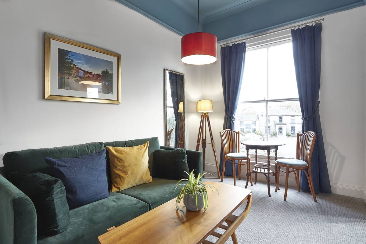 A living room with a green sofa, a wooden coffee table, a framed picture on the wall, a red pendant light, and a view of the city through large windows.