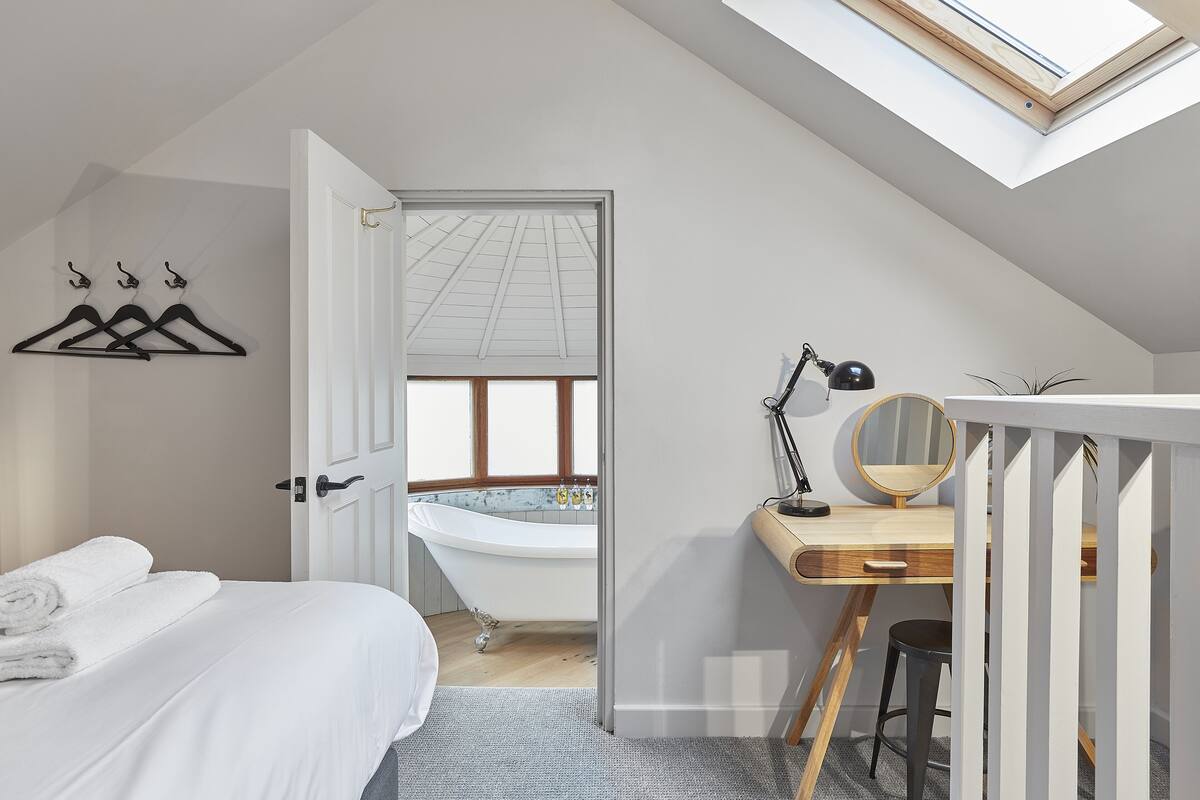 A bedroom with a skylight, a bathtub, a desk with a mirror, and a bed with towels.