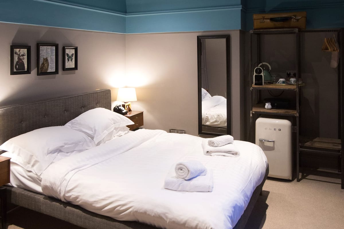A neatly made bed with white linens, a bedside table with a lamp, a small refrigerator, and a mirror on the wall.