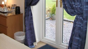 Habitación de hotel con cama, escritorio con lámpara, cafetera y vista a un jardín por una puerta de vidrio.