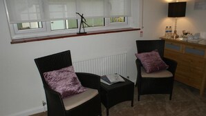Una habitación con dos sillones de mimbre con cojines, una mesita con una revista, un radiador, una ventana con persianas y una lámpara sobre un aparador.