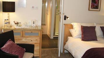 A hotel room with a bed, a chair, a desk with a lamp, a coffee maker, and a shower area.