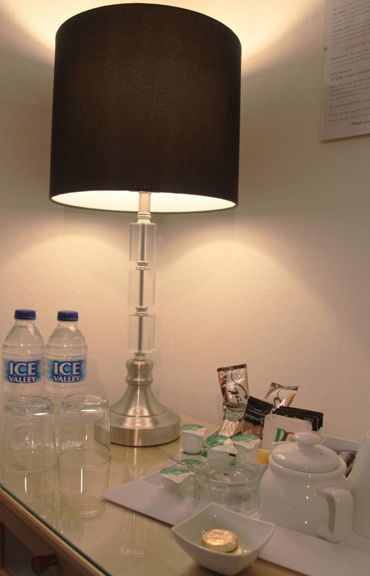 A hotel room with a table lamp, two bottles of water, a coffee maker, and some toiletries.