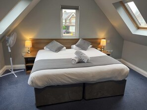 A bedroom with a large bed, two bedside tables, a standing fan, and a skylight. - The Corner House Hotel Gatwick (Horley)