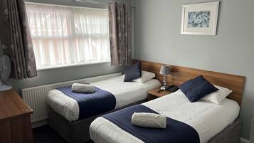 A hotel room with two beds, a bedside table with a lamp, a framed picture on the wall, and a window with curtains.