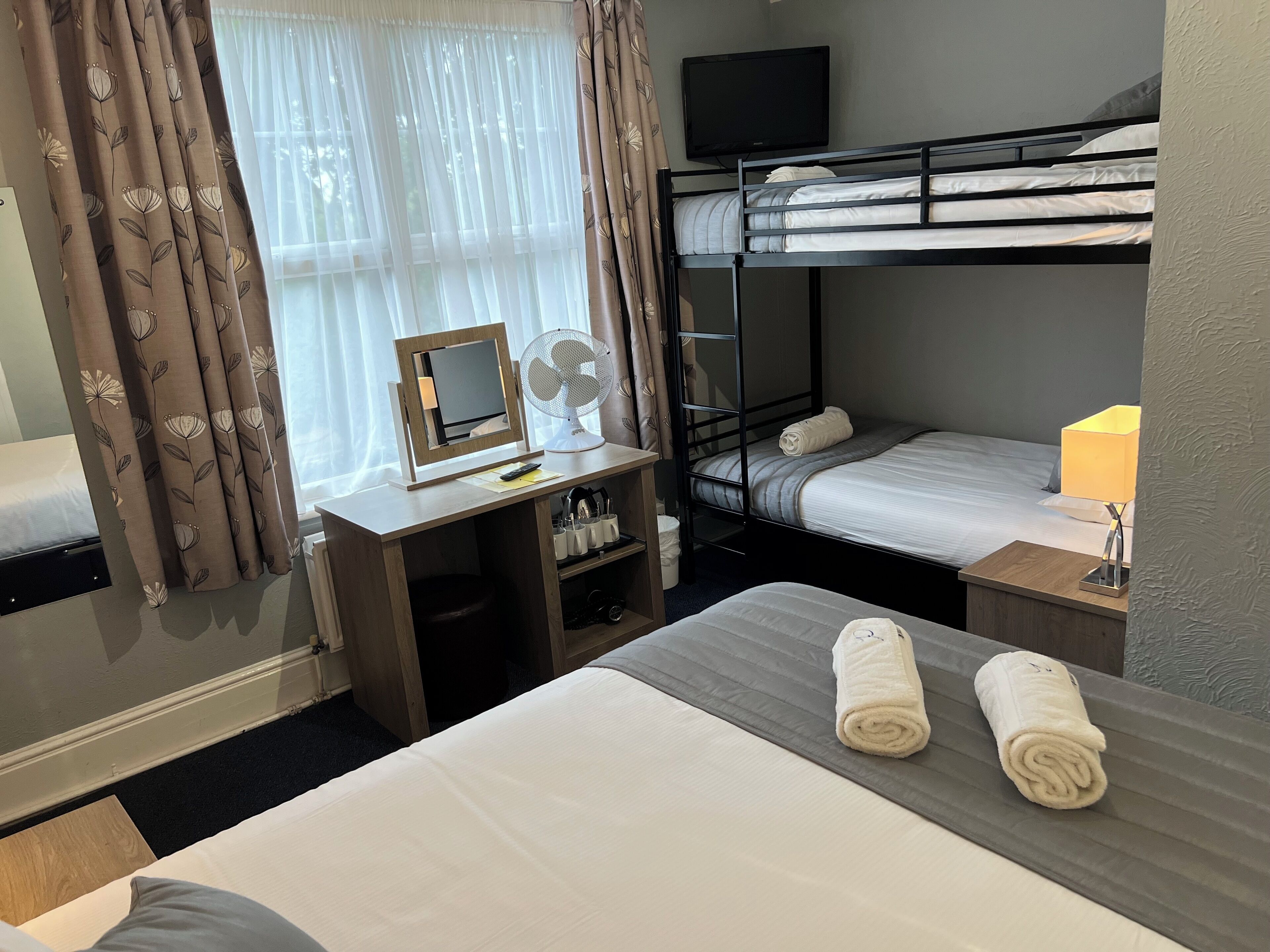 Ein Hotelzimmer mit Etagenbett, Schreibtisch mit Spiegel, Ventilator und Nachttisch mit Lampe.