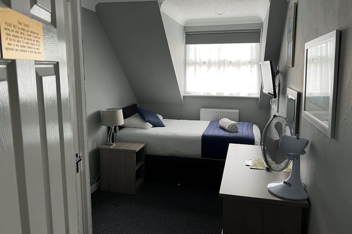 Pieni hotellihuone, jossa on sänky, yöpöytä, lamppu, tuuletin ja ikkuna, jossa on verhot.