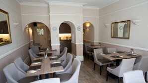 A dining area with multiple tables and chairs, a kitchen in the background, and a mirror on the wall. - The Corner House Hotel Gatwick (Horley)
