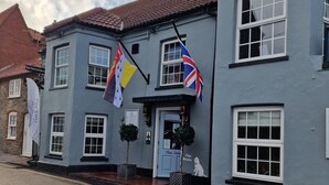A two-story building with a blue facade, multiple windows, and a sign reading "The Two Lifeblood." Flags are displayed outside, and there are potted plants on the ground. - The Two Lifeboats (Sheringham)