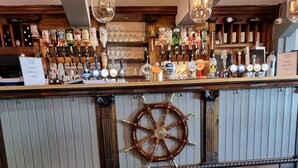 A bar area with a dog sitting on the floor, a ship's wheel decoration, and a variety of bottles and glasses on the shelves. - The Two Lifeboats (Sheringham)