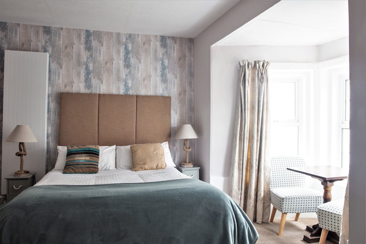 A bedroom with a large bed, two bedside tables with lamps, a patterned headboard, and a view of a dining area with a table and chairs.