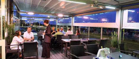 An outdoor dining area with tables and chairs, a waiter, and a view of the cityscape through large windows.