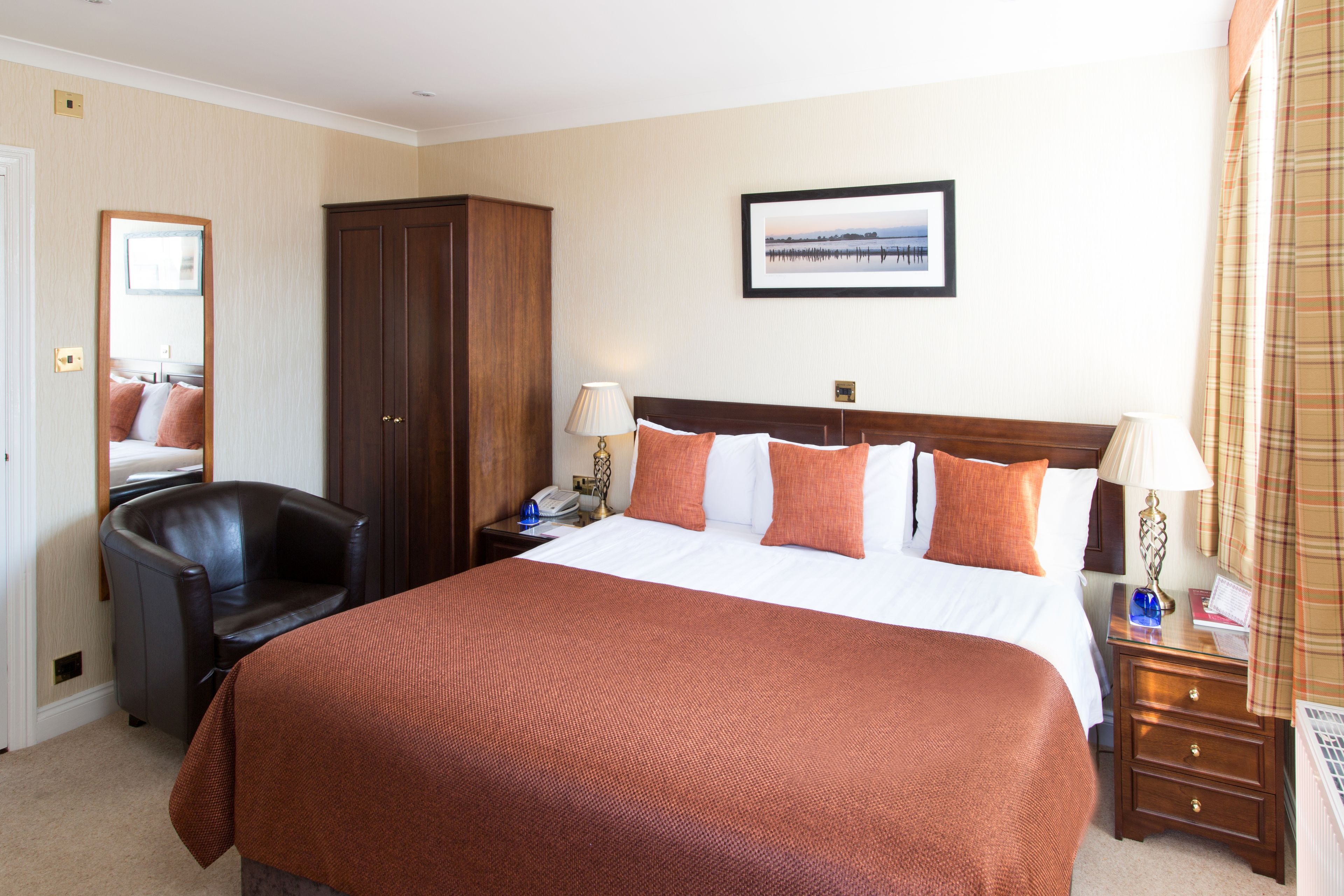 A hotel room with a large bed, a wooden wardrobe, a chair, a bedside table with lamps, and a framed picture on the wall.