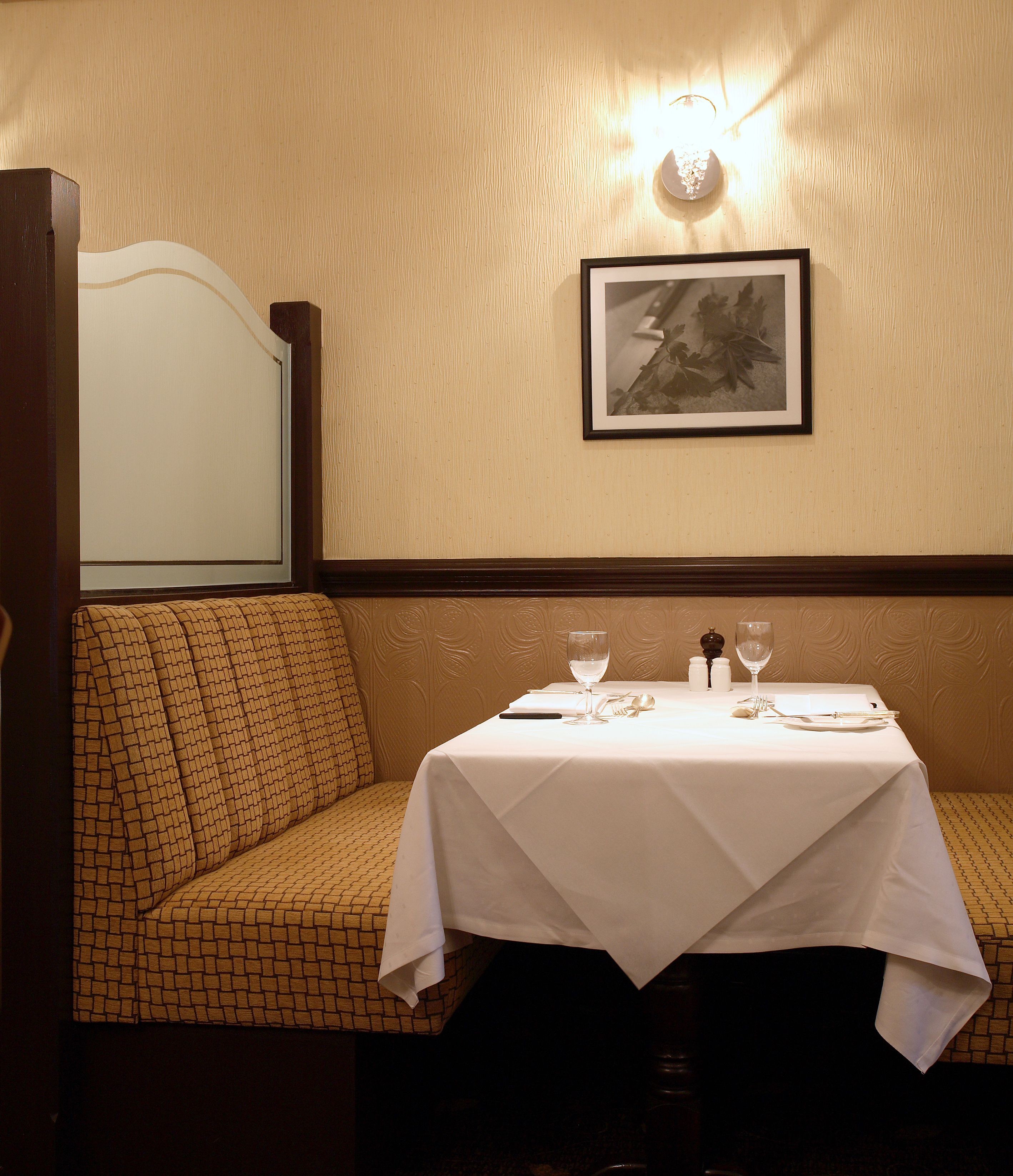 A dining area with a table set for two, a cushioned bench, a framed picture on the wall, and a wall-mounted light fixture.