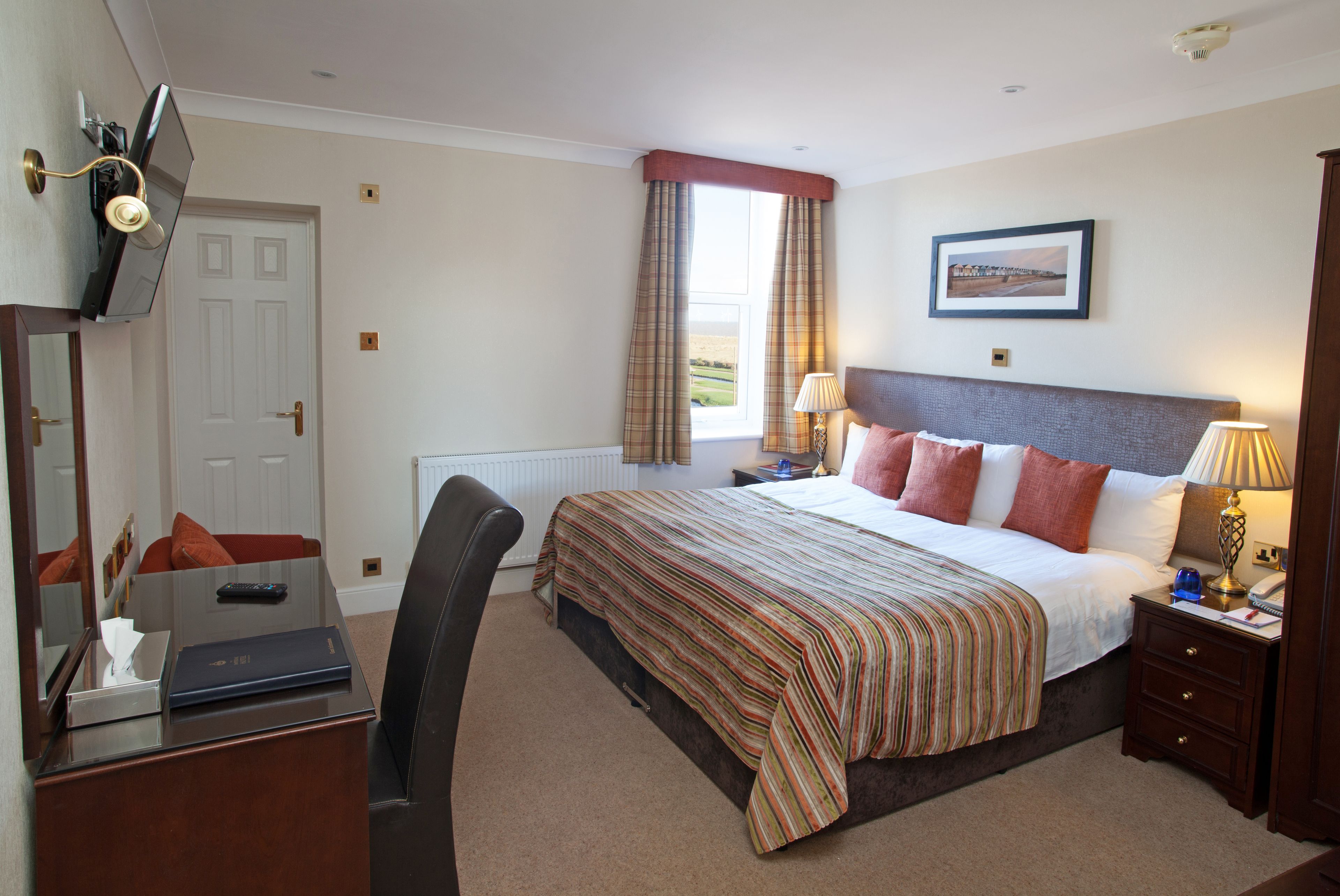 A hotel room with a bed, a desk with a computer, a chair, a nightstand with a lamp, and a window with curtains.