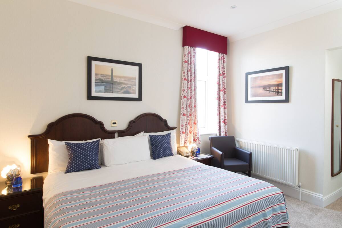 Une chambre d’hôtel bien rangée, avec un grand lit, deux tableaux encadrés aux murs, une fenêtre avec des rideaux, une chaise et une table de chevet avec une lampe.