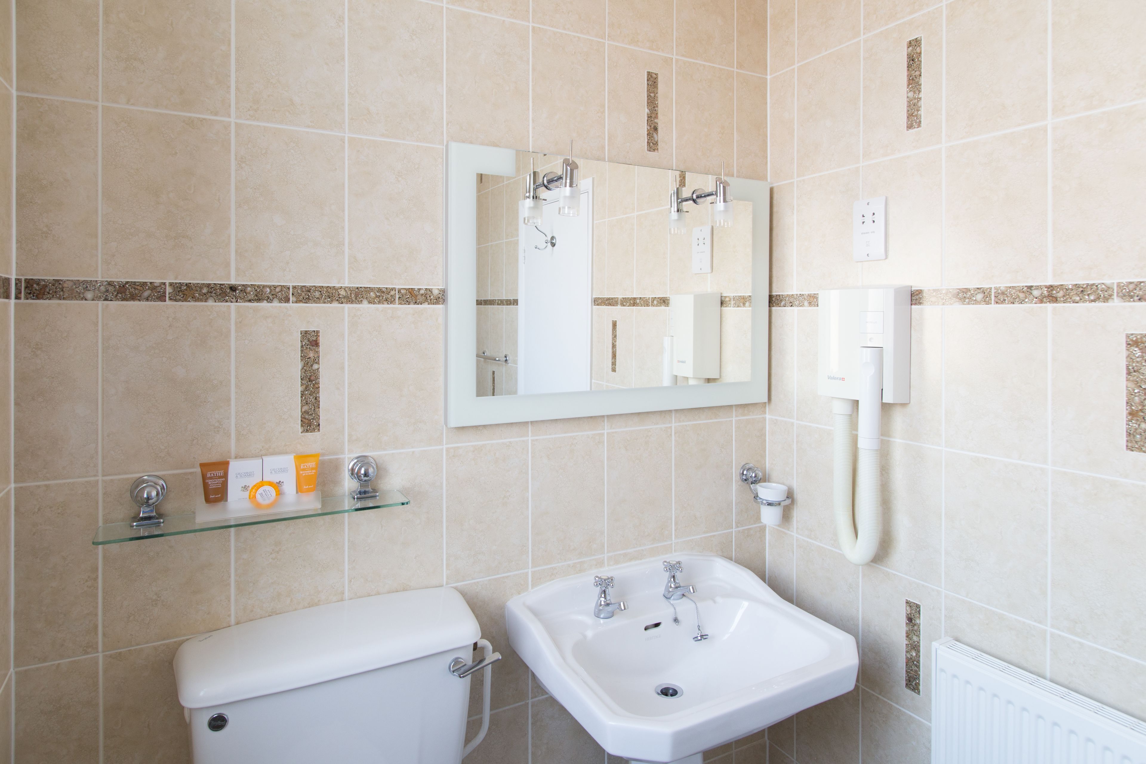 A bathroom with a toilet, sink, and mirror. There is a shelf with toiletries and a towel rack.