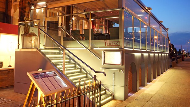Restaurante com terraço envidraçado e mesas ao ar livre, escadaria que leva à entrada e placa com o nome 'The Terrace'.