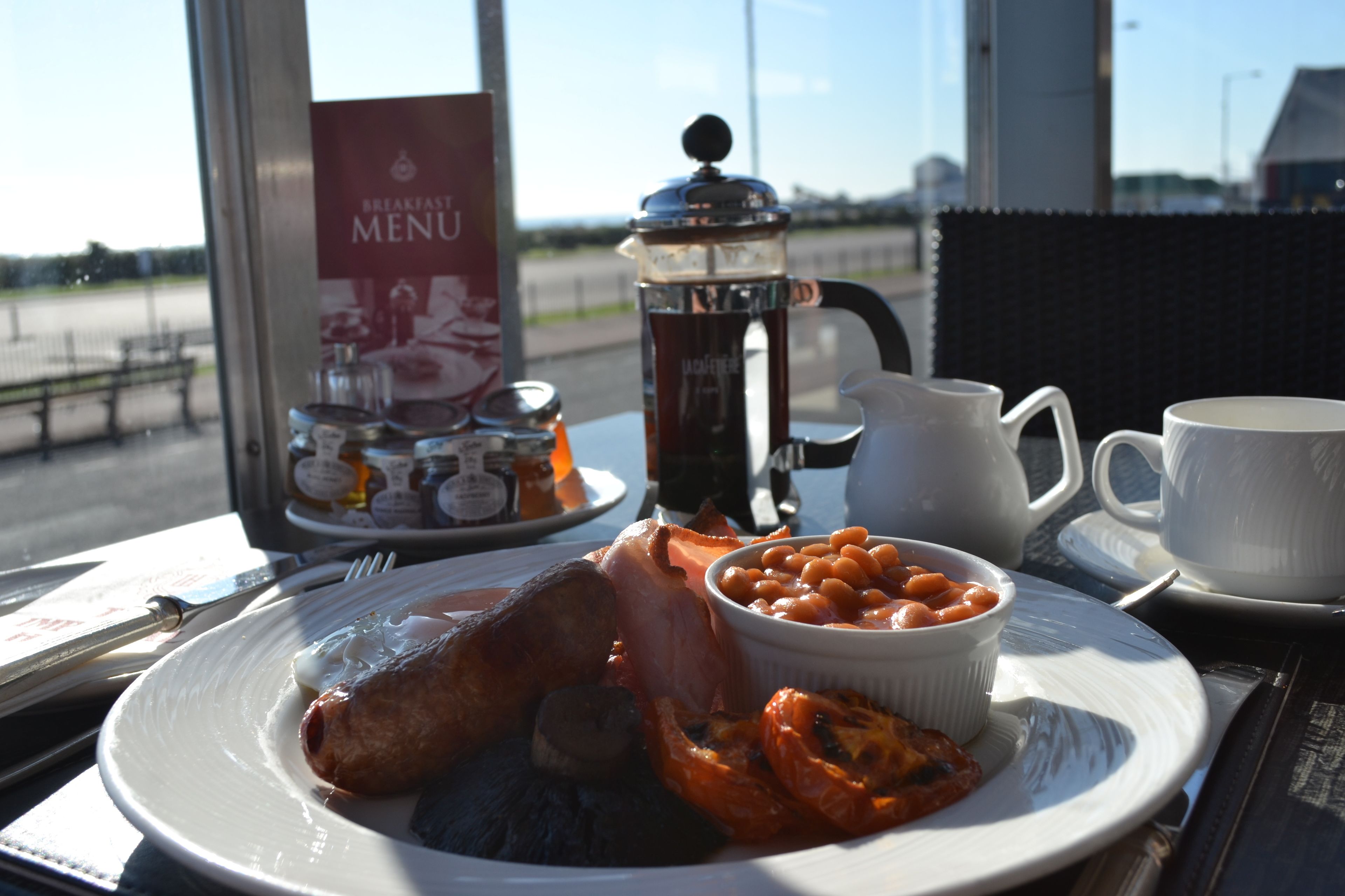 A breakfast plate with ham, beans, and tomatoes, a coffee pot, and a menu on a table by a window.