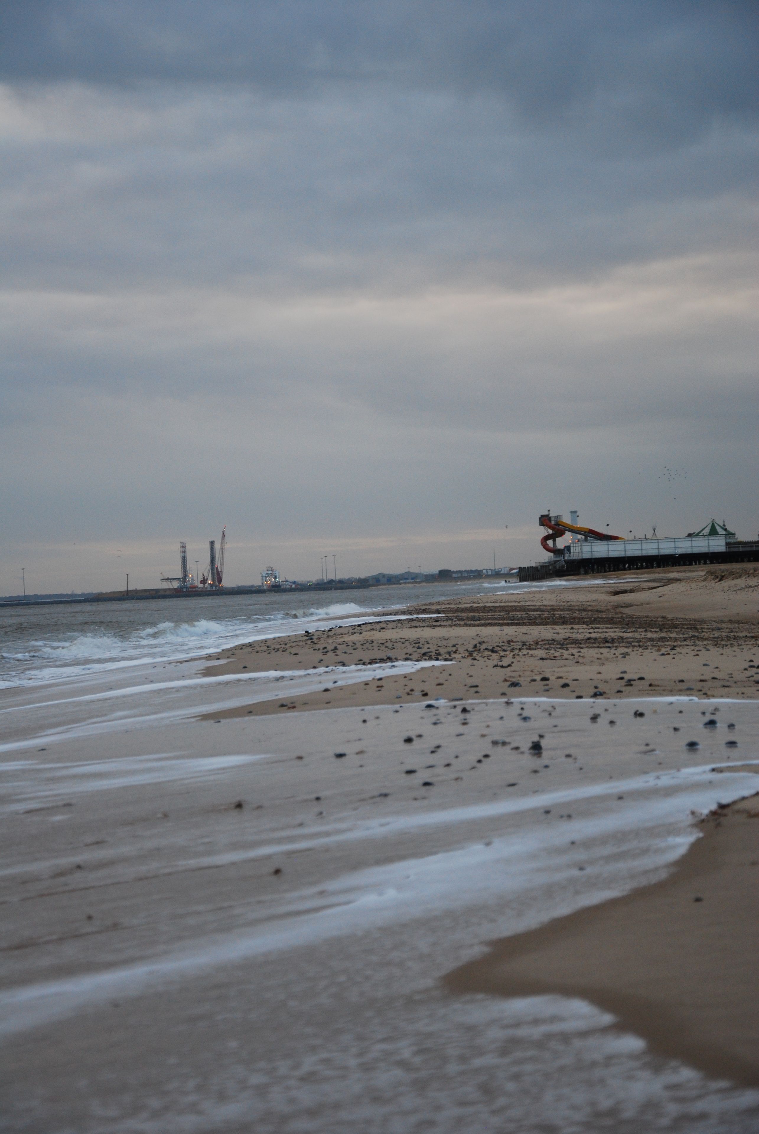 A beach with waves, a pier, and industrial structures in the distance.