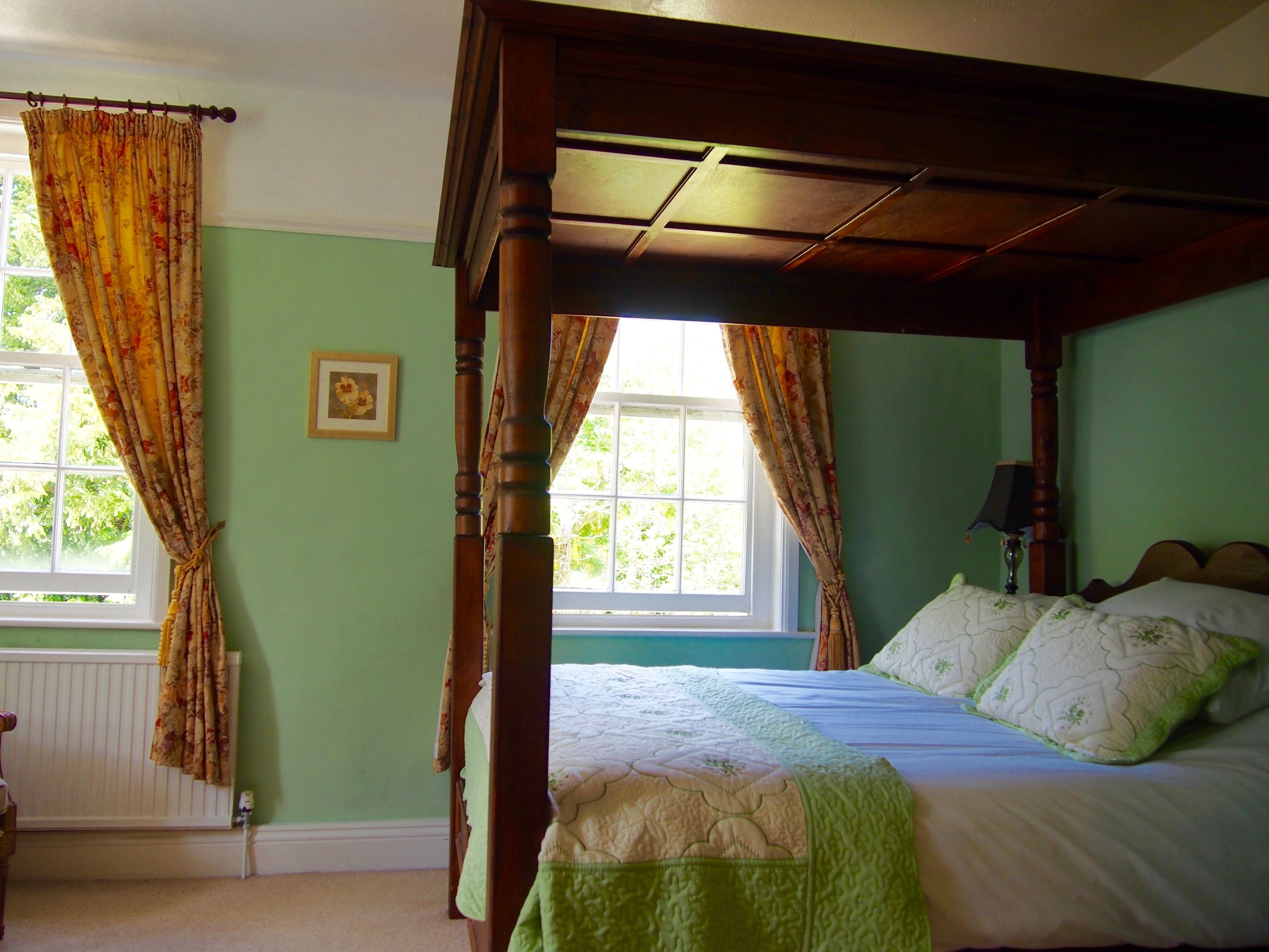 A four-poster bed with a green quilt, a bedside table with a lamp, and a window with curtains.