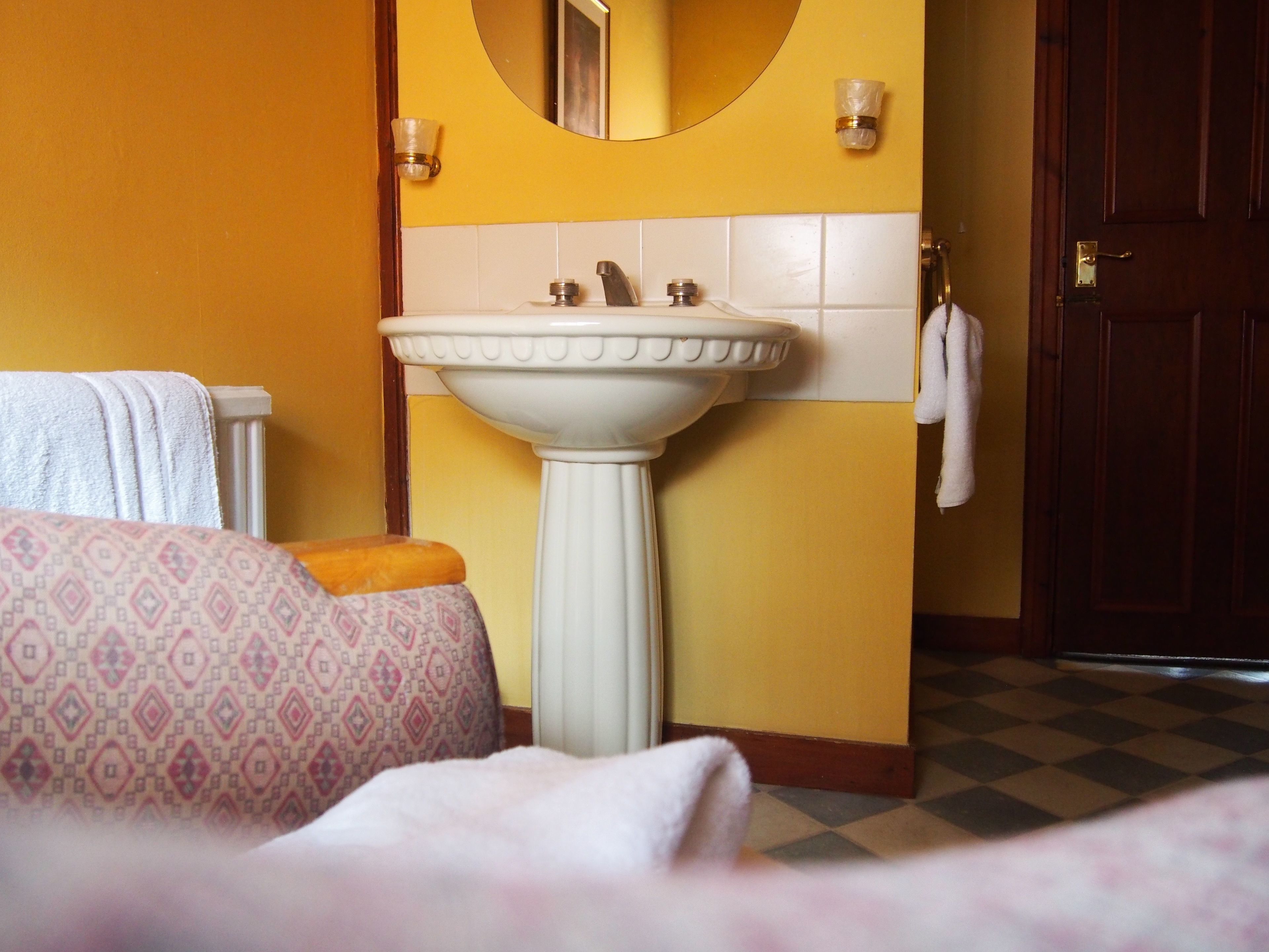 Un bagno con un lavandino bianco su piedistallo, pareti gialle, un tappetino da bagno fantasia e un pavimento a scacchi.
