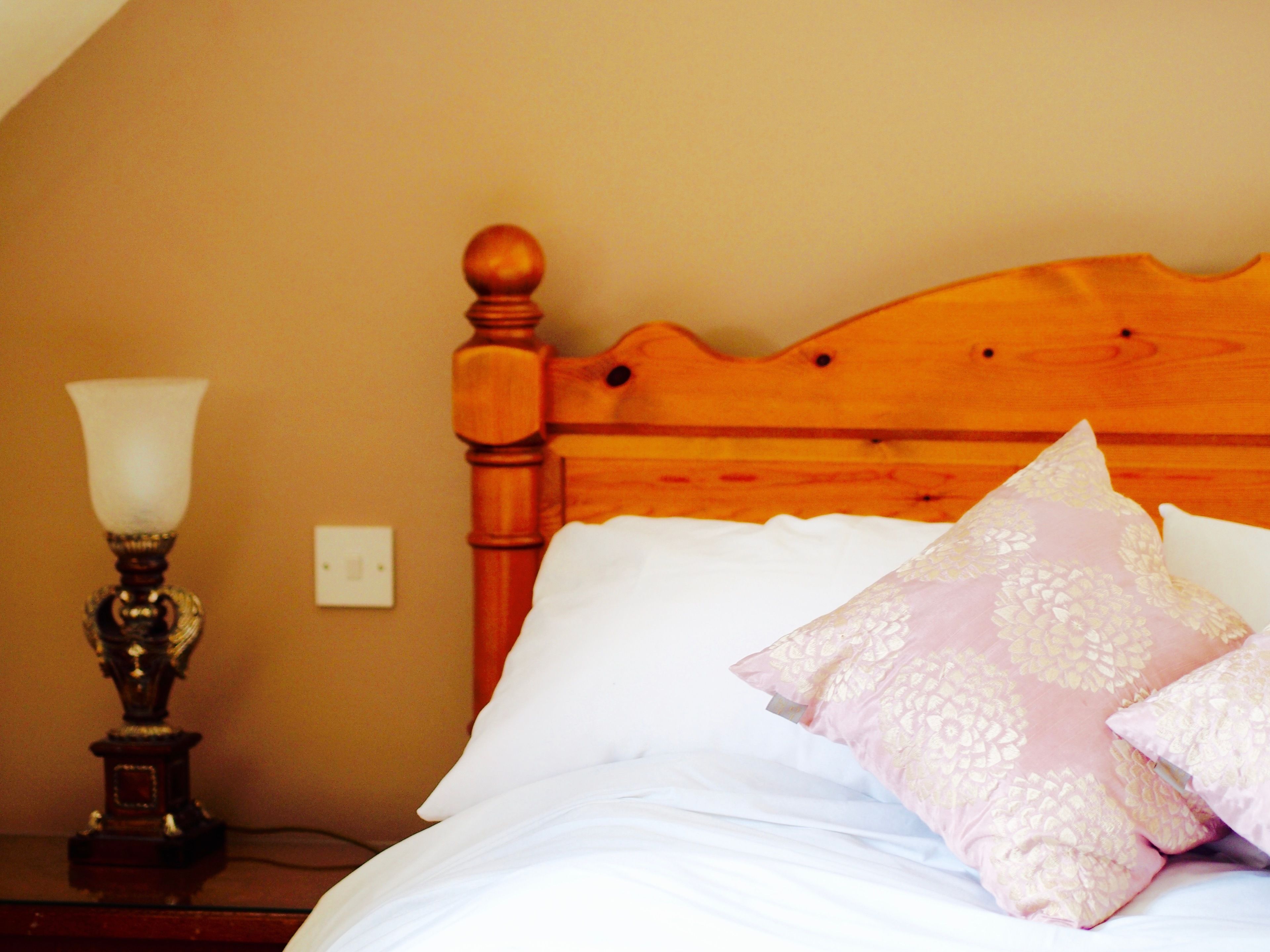 A wooden bed with white linens and decorative pillows, a bedside table with a lamp, and a wall socket.