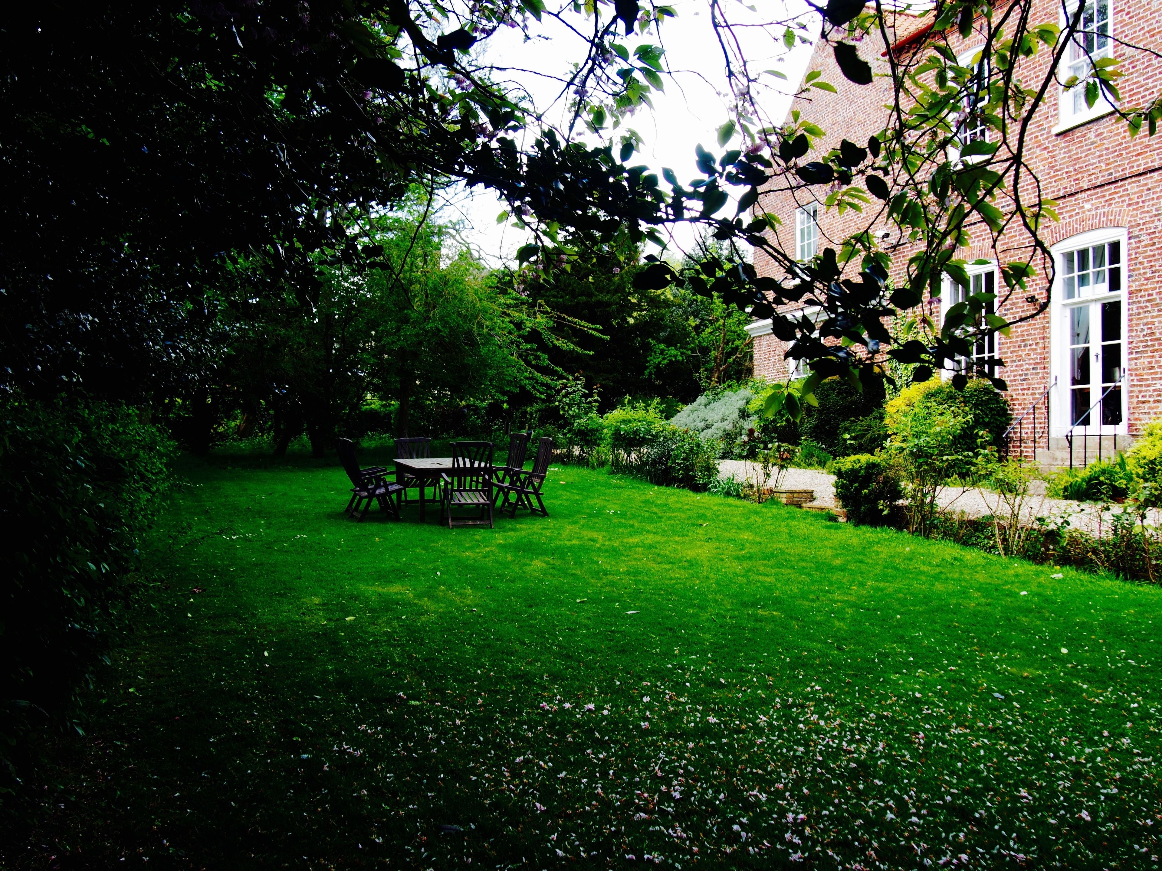 A well-maintained garden with a wooden table and chairs, surrounded by lush greenery and a brick building in the background.