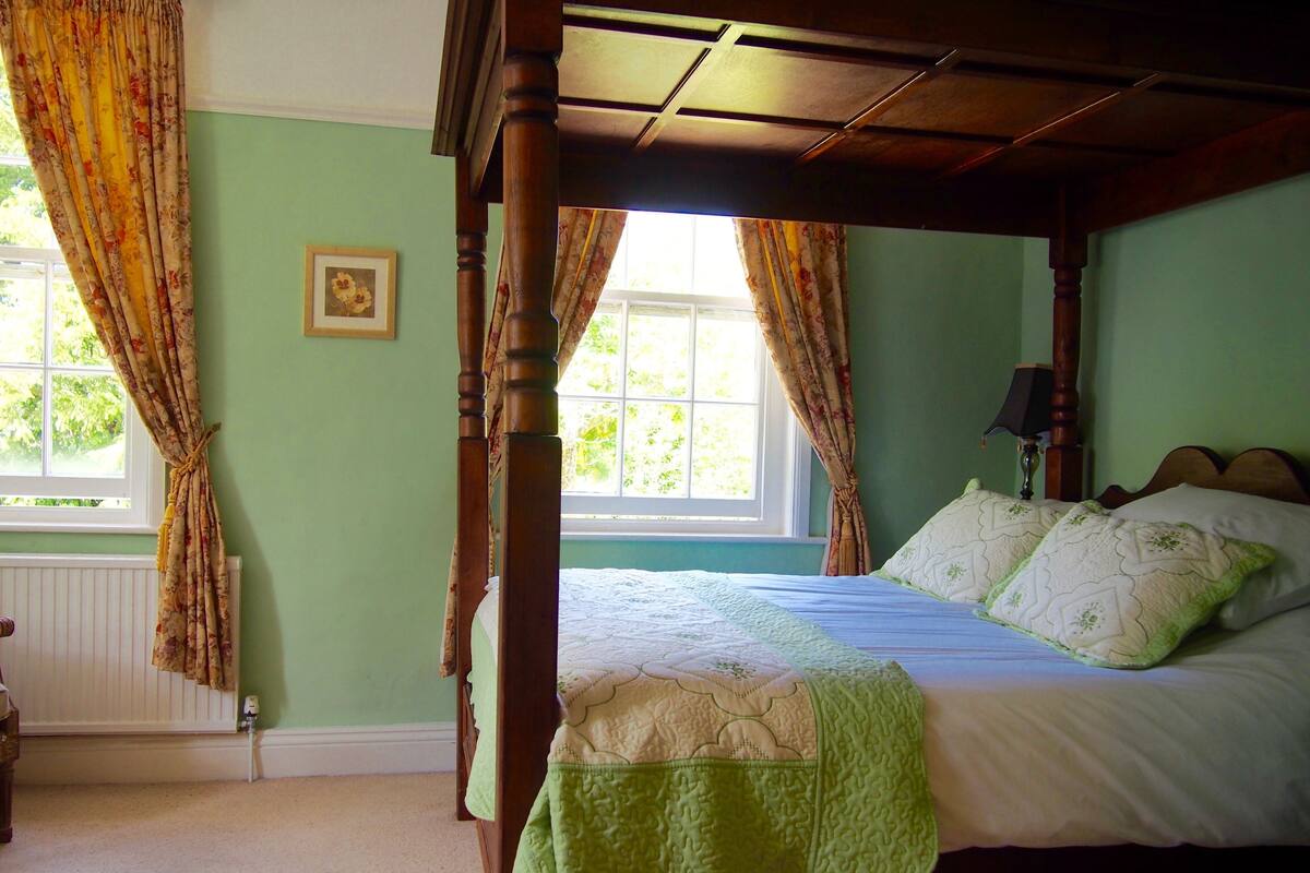 A four-poster bed with a green quilt, a window with floral curtains, and a framed picture on the wall.
