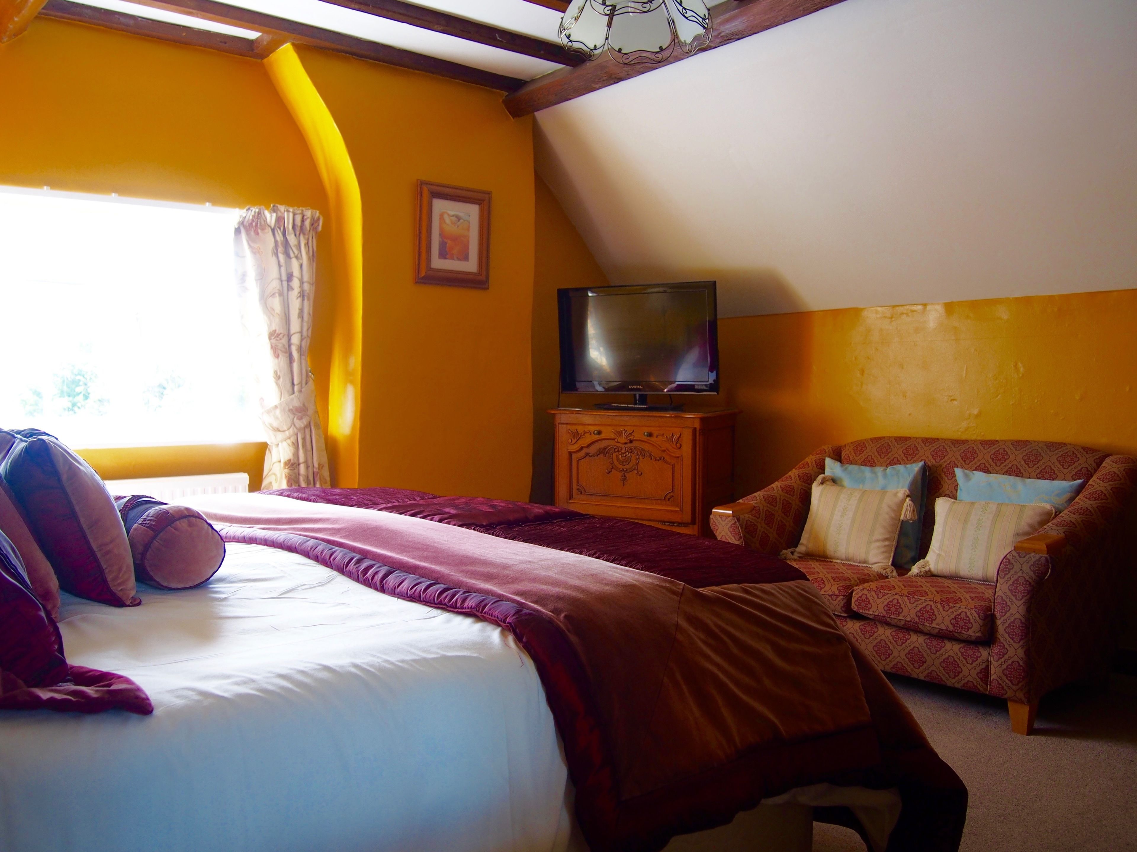 A bedroom with a bed, a television on a wooden cabinet, a patterned armchair, and a window with curtains.