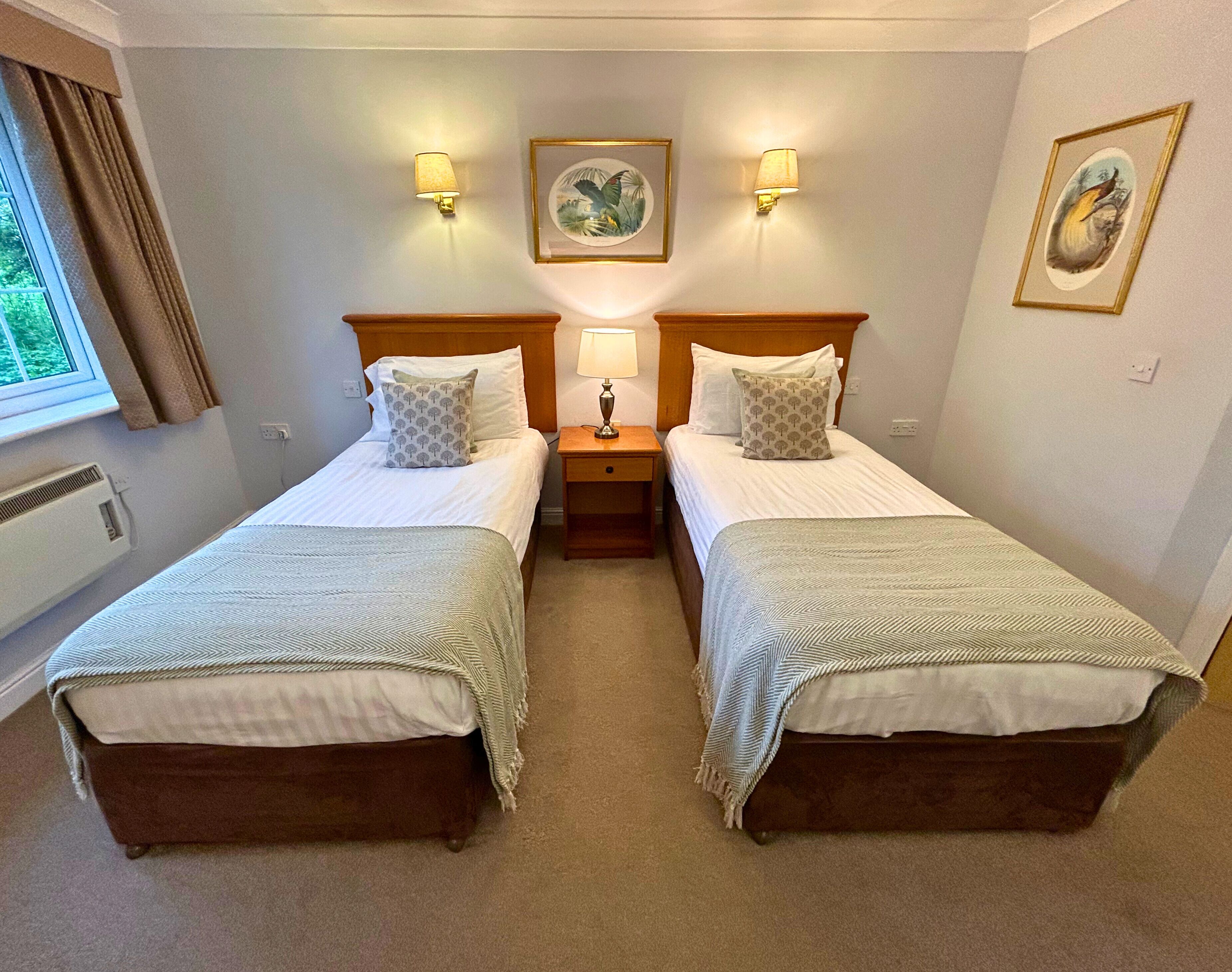A twin room with two beds, a nightstand between them, a window with curtains, and wall-mounted lamps.