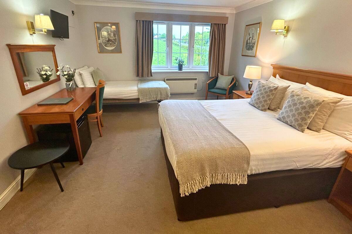 A hotel room with a large bed, a desk with a chair, a small table, a mirror, and two wall-mounted lamps.