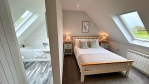 A bedroom with a skylight, a bed, bedside tables with lamps, a view of the outside, and a bathroom with a sink and bathtub. - New Forest Lodge (Salisbury)